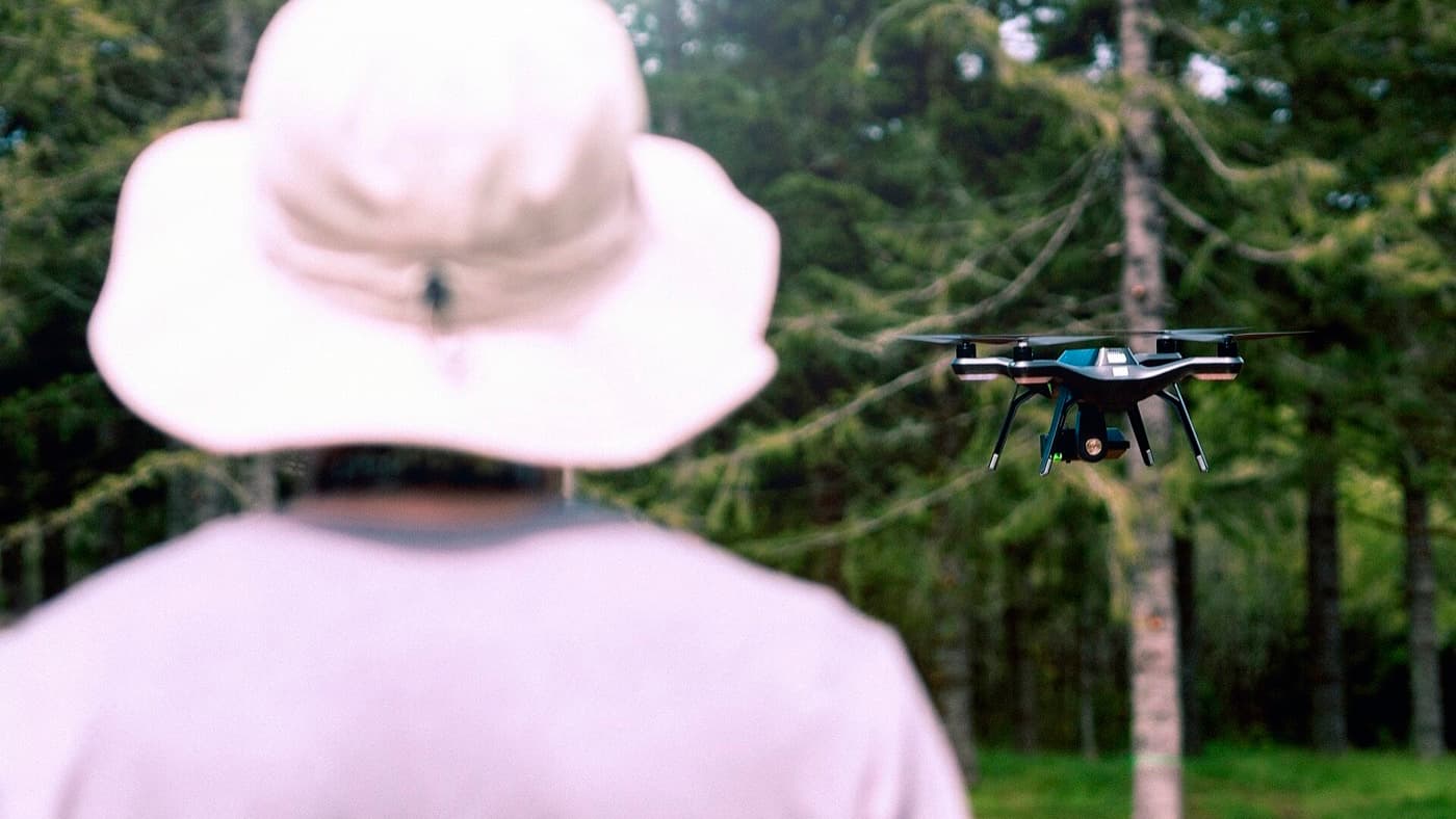 A camera drone hovering with its gimbal-mounted camera visible, an operator holding the controller in the background