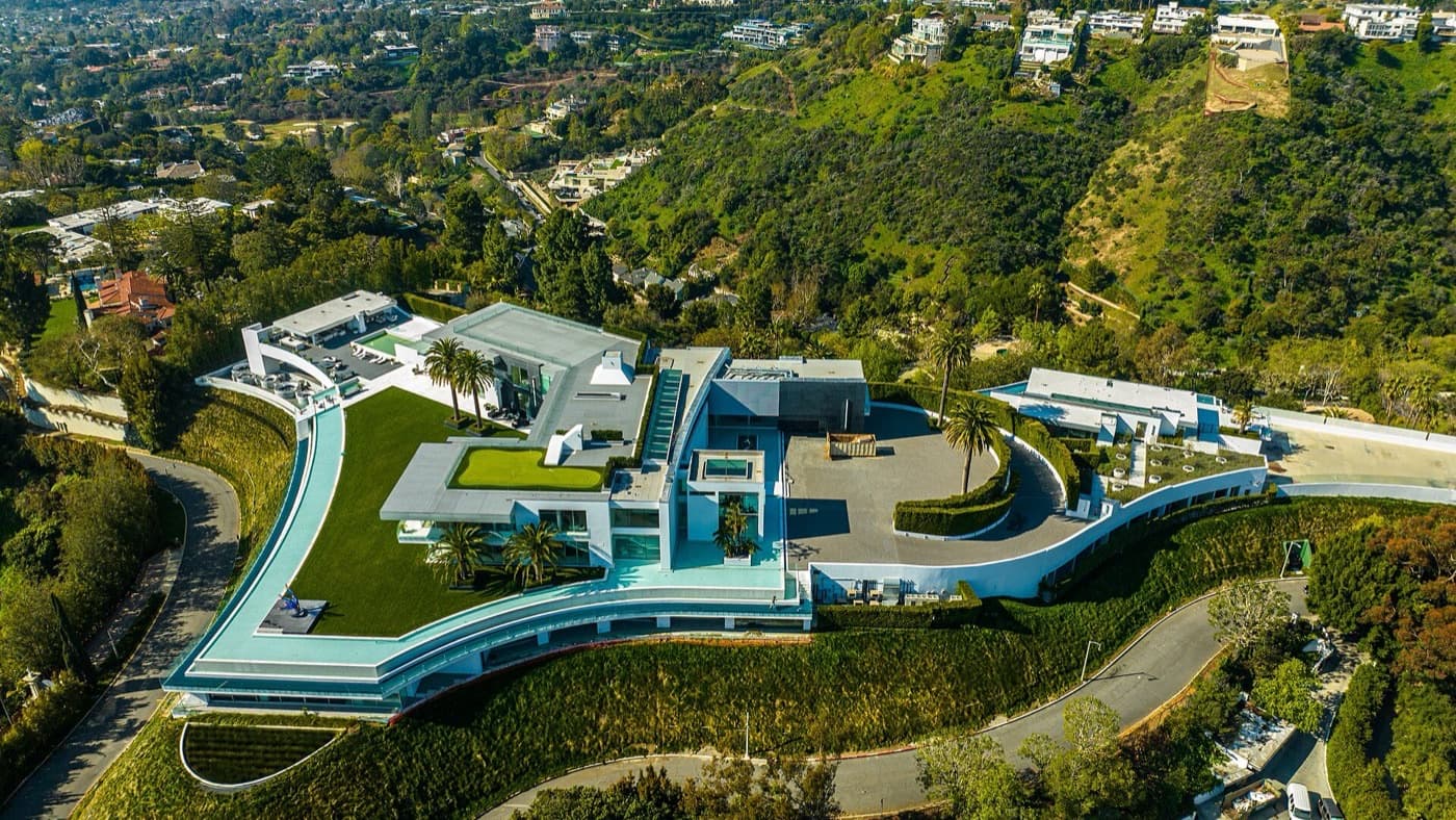 Aerial view of a large modern hillside estate showing its full footprint, grounds, pool terraces and the city beyond