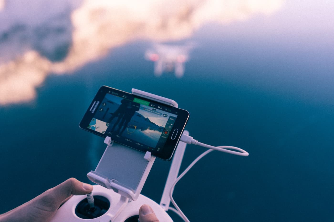 Mâini pe telecomanda unei drone, cu un telefon care arată imaginea camerei și drona deasupra apei