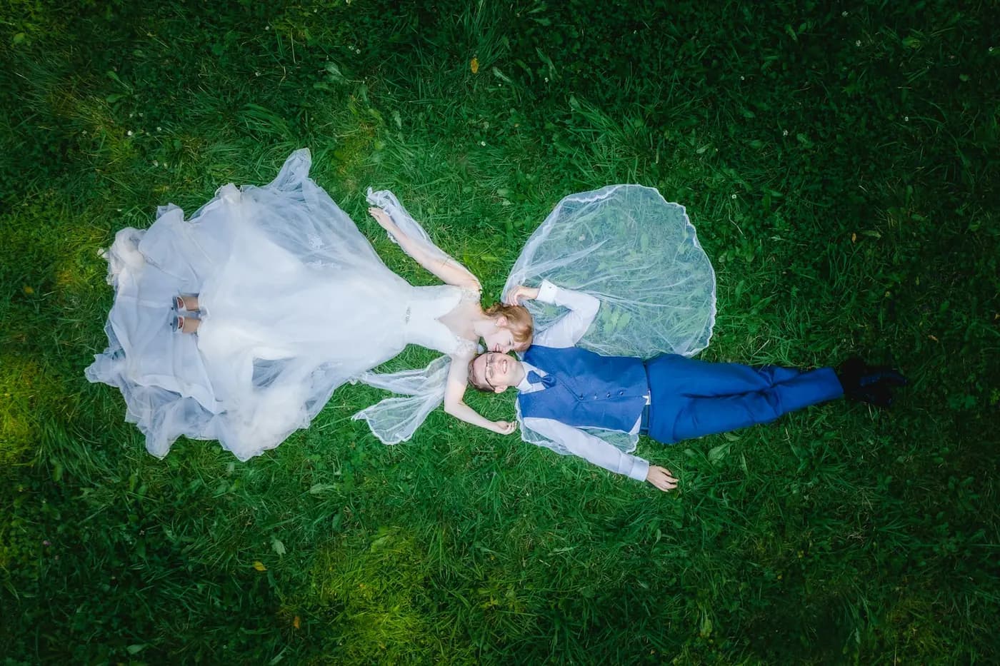 Overhead view of a bride and groom lying on a lawn, her veil spread around them